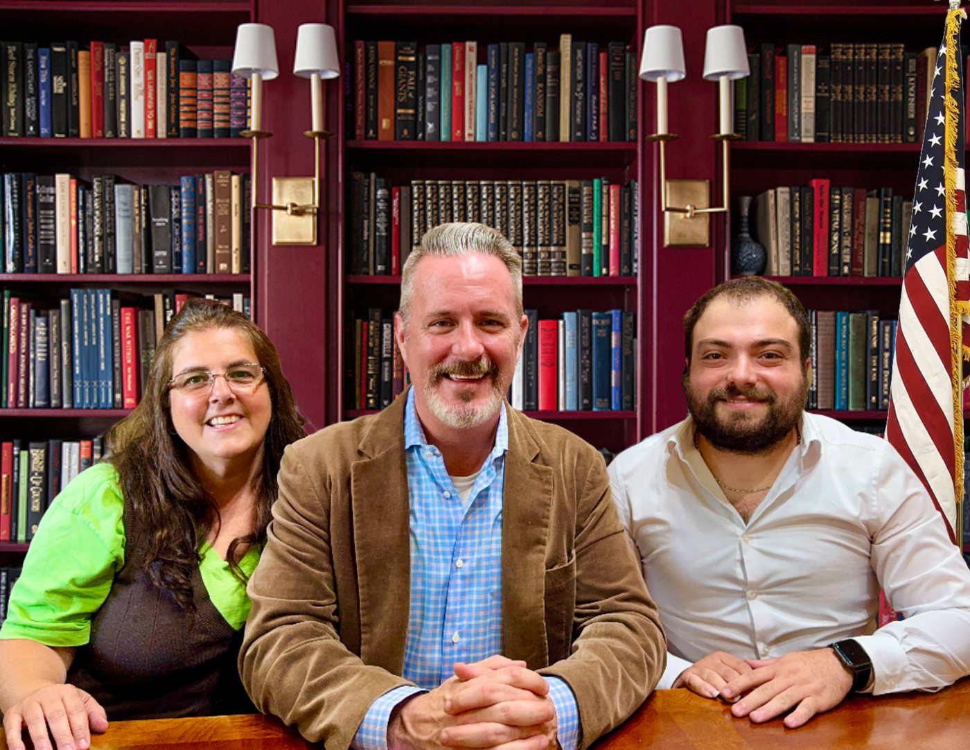 Three members of the Shea Law, LLC team smiling at a desk in front of a library bookcase and an American flag